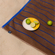 Brown beach towel with blue lines featuring a plate of limes on a beach setting.