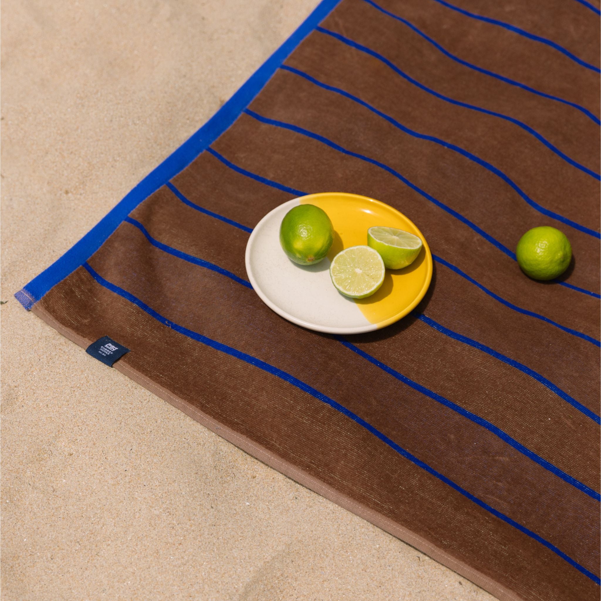 Brown beach towel with blue lines featuring a plate of limes on a beach setting.