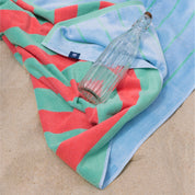 A beach towel with green, coral, and blue stripes laid out on sand with a clear water bottle resting on top.