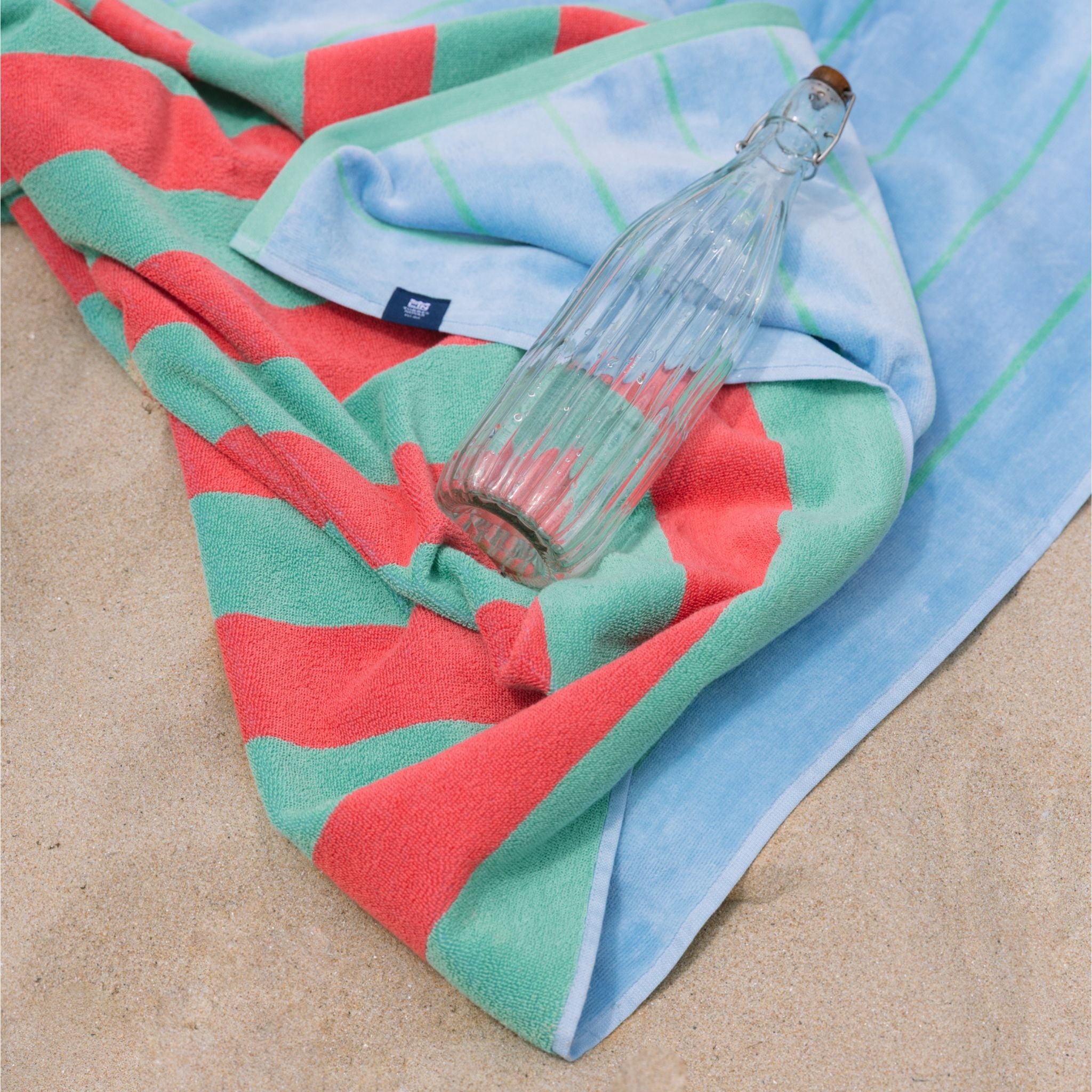 A beach towel with green, coral, and blue stripes laid out on sand with a clear water bottle resting on top.