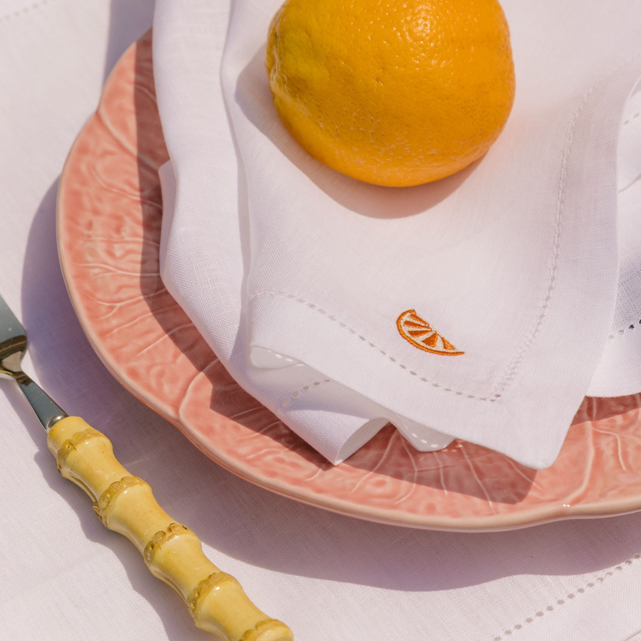 Pink plate with a white napkin featuring a logo, an orange, and a yellow-handled fork on a light background.