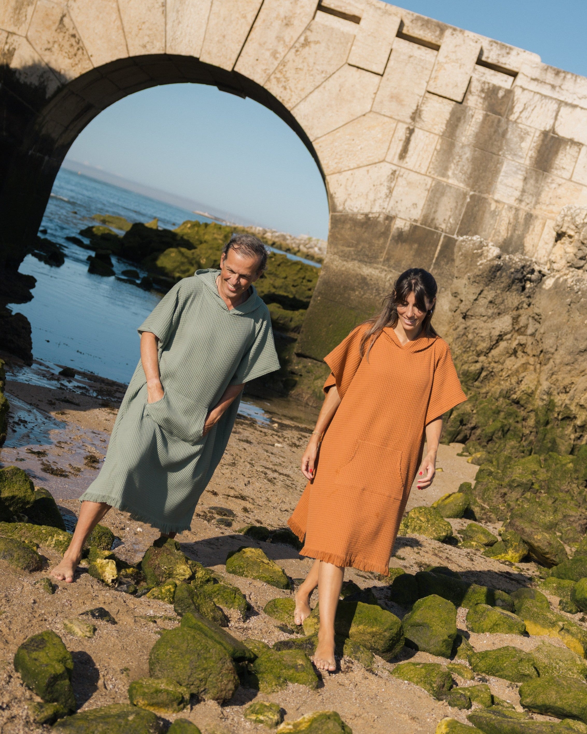 Two models wearing loose, short-sleeve beach ponchos walk barefoot on a rocky beach under a large stone archway. One poncho is sage green and the other is terracotta orange, set against a natural coastal background.